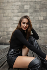 Beautiful happy stylish elegant woman in fashion trendy black long sleeve sweater, leather shorts and high shoes sitting on wooden bench near vintage brick wall on the street