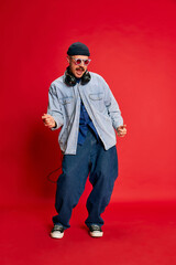 Portrait of man in stylish modern clothes, oversized jeans, shirt and hat posing in headphones over red background. Concept of modern fashion, lifestyle, music culture