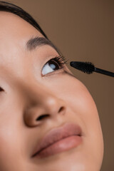Close up view of asian woman applying mascara isolated on brown.