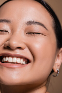 Cropped view of smiling asian woman closing eyes isolated on brown.