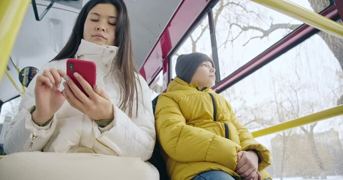 Front view of passengers sitting on bus, waiting. Brunette girl smartphone holding, texting, chatting, scrolling, small boy looking in window. Concept of public transport.