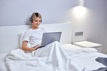 beautiful caucasian woman during online dating of video meeting with someone, smiling, in headphones, having rest relaxing on bed at home. woman use modern laptop. modern technologies, quarantine