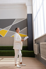 full length of multiracial businesswoman in white suit standing with papers and smiling at camera in office.