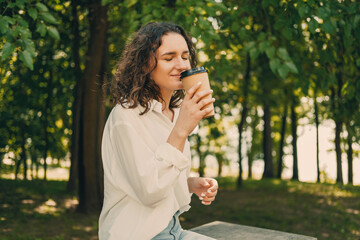 Fototapeta premium Young curly woman enjoys to go coffee with eyes closed in a green park.