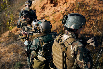 Obraz premium Squad of Young Athlete Fully Equipped Friendly Soldiers in Camouflage on a Reconnaissance Military Mission, Aiming Rifles. Rear View On Men Moving in Formation Through Mountains.