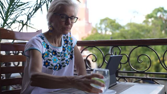 Happy senior mature woman types on laptop tablet computer pauses to drink, think and contemplate, wearing ethnic clothes sitting at a balcony cafe as traffic passes below. Concept of digital nomad.