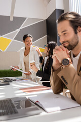 cheerful multiethnic businesswomen talking near young manager looking at laptop on blurred foreground.