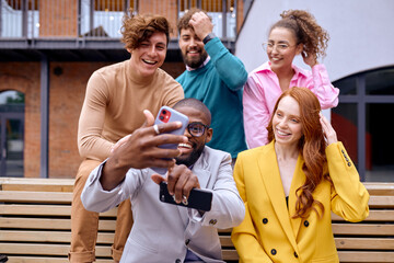 Excited diverse colleagues have fun taking photo on smartphone, smiling, men and women after successful cooperation meeting, dressed in fashionable stylish clothes, sitting on bench outdoors