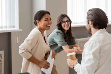 joyful interracial businesswomen laughing near young manager standing with coffee to go.