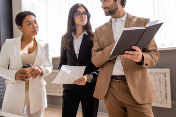 young manager showing notebook to multicultural businesswoman standing with paper and takeaway drink.