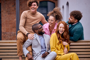 happy business executives talking in city park while taking coffee break on bench, having fun, laughing, talking jokes. attractive ladies and guys relaxing together, sitting on bench