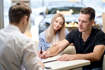 young male shop assistant explaining talking offering buying new car to young caucasian family husband and wife at auto dealer shop, sit talking discussing. in cars showroom