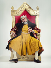 King, royalty and bored man on throne in studio isolated on gray background. Monarch, medieval royal and annoyed senior male, leader or frustrated ruler, looking at hand and sitting on golden seat.