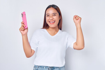 Beautiful excited Asian girl wearing casual white t-shirt holding mobile phone, raising her fist,...
