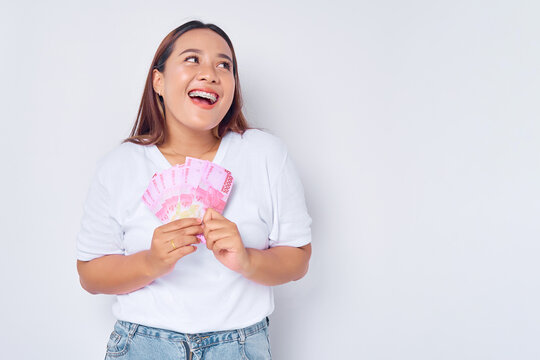 Excited young blonde woman girl Asian wearing casual white t-shirt holding money rupiah banknotes and looking at empt space isolated on white background. Profit and wealth concept