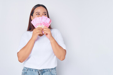 Smiling young blonde woman girl Asian wearing casual white t-shirt covering her face with money rupiah banknotes and looking aside isolated on white background. people lifestyle concept