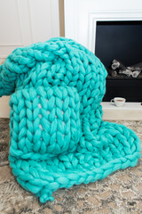 A warm plaid knitted from merino wool of a beautiful aquamarine shade lies near the fireplace along with a cup of tea. The concept of comfort.