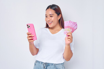 Smiling young blonde woman girl Asian wearing casual white t-shirt using mobile phone and holding money rupiah banknotes isolated on white background. Fast credits concept