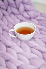 The concept of coziness and comfort is a pink-lavender knitted blanket with a white cup of hot tea on it. Place for inscription, vertical photo.