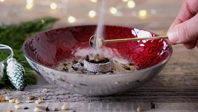 Burning aromatic incense resin (boswellia, frankincense), styrax and rose blossom petals in a red incense bowl. A hand is putting the incense with a brass spoon on the glowing incense charcoal. 