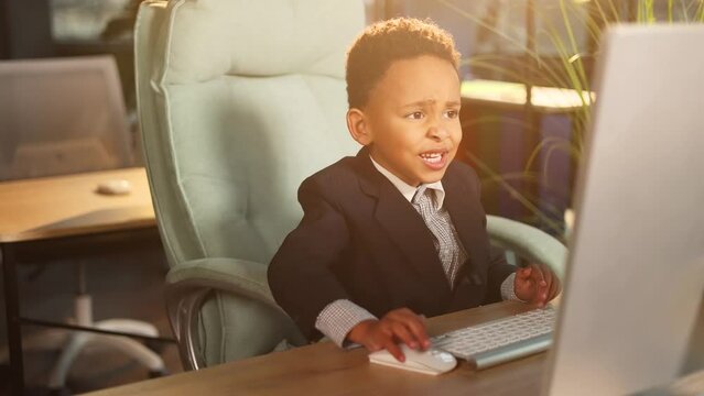 Cute successful little businessman child kid looking at computer screen browsing typing in the modern office workplace Ambitious young people or early start of business concept