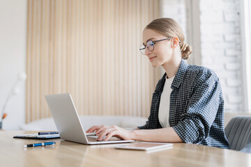 A smiling business woman in glasses and a shirt, using a laptop in the office, typing text on the keyboard.