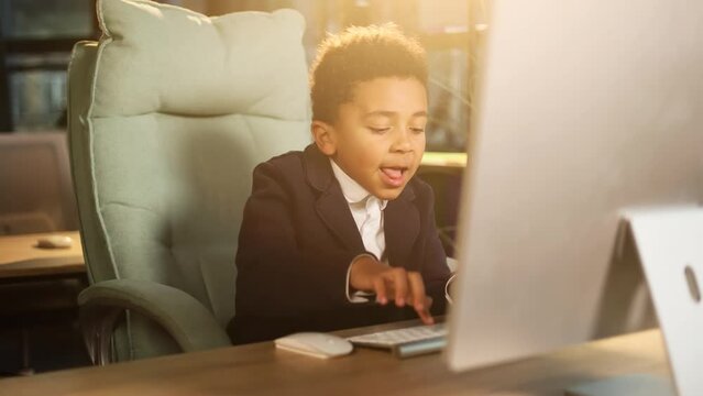 Cute successful little businessman child kid looking at computer screen browsing typing in the modern office workplace Ambitious young people or early start of business concept