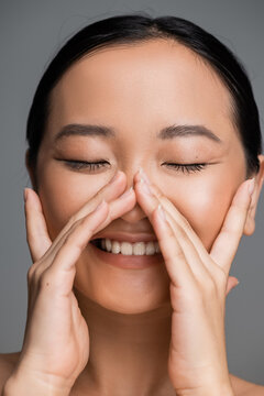 young asian woman with closed eyes and nude makeup smiling and touching face isolated on grey.