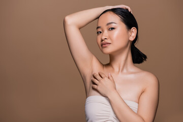 Pretty asian woman with naked shoulders touching fair and looking at camera isolated on brown.