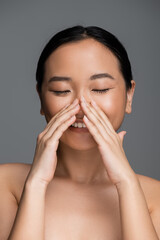 happy asian woman with closed eyes and perfect skin touching face isolated on grey.