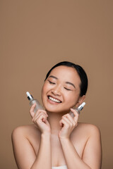 Pleased asian woman holding cosmetic serums isolated on brown.