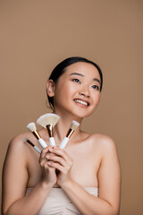 Positive asian model in top holding cosmetic brushes isolated on brown.