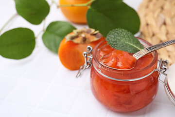 Jar and spoon of tasty persimmon jam, ingredients on white tiled table. Space for text