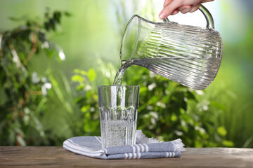 Woman pouring water from jug into glass on wooden table outdoors, closeup