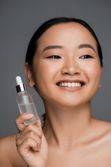 young and happy asian woman with clean skin holding bottle of cosmetic serum isolated on grey.