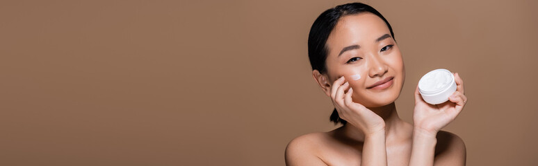 Smiling asian model applying face cream on cheek isolated on brown, banner.