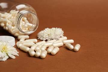 Open medicine bottle, scattered pills and flowers on brown background, space for text