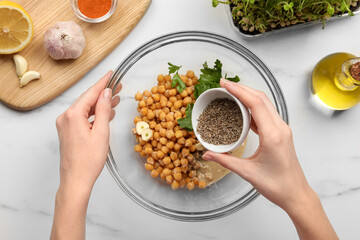 Woman adding cumin into bowl with ingredients for delicious hummus at white marble table, top view