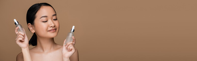 Brunette asian woman with naked shoulders holding bottles of cosmetic serums isolated on brown,...