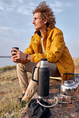 handsome male drinking tea in nature sitting on top of mountain, relaxing during camping,hiking, trekking. relaxed caucasian guy in yellow casual coat enjoy the landscape, wonderful nature view.