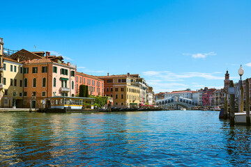 Fototapeta na wymiar Beautiful cityscape in Venice, Italy. Picturesque old buildings surround the iconic Rialto Bridge on the Grand Canal, accompanied by gondolas and boats under vivid blue sky on sunny spring day.