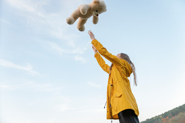 Young woman toss up soft toy outdoors. Beautiful girl in yellow jacket with teddy bear on sky...