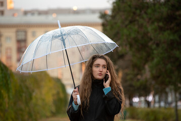 Girl walking in the park and chat. Smiling Young woman talking on phone in autumn park with transparent umbrella