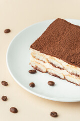 Homemade tiramisu cake on white plate against beige background surrounded by coffee beans . Italian dessert made of savoiardi cookies filled with mascarpone cheese and sprinkled with cocoa powder