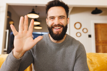 Portrait of happy biracial bearded young man waving hand while sitting on sofa in living room - Powered by Adobe