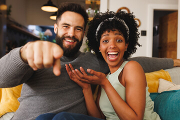 Portrait of cheerful biracial couple laughing and gesturing while sitting on sofa in living room - Powered by Adobe