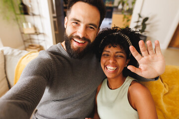 Obraz premium Portrait of happy biracial bearded man waving hand and taking selfie with girlfriend on sofa