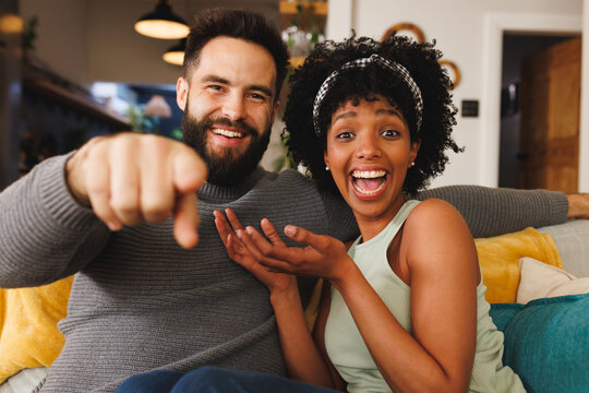 Portrait of cheerful biracial couple laughing and gesturing while sitting on sofa in living room