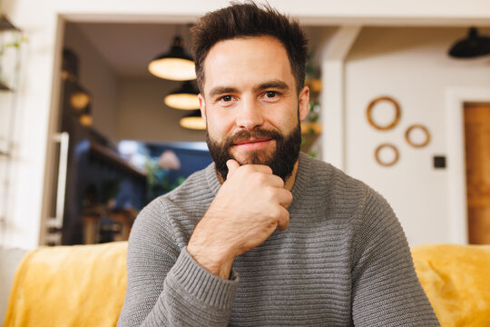 Portrait of biracial bearded young man with hand on chin smiling and sitting on sofa in living room