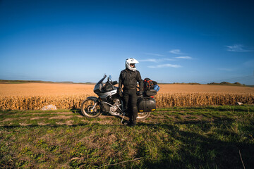 Obraz premium A man, a motorcycle driver, stands next to his touring motorcycle. Large side bags for luggage. A field of dry yellowed corn. Freedom and travel transportation concept.